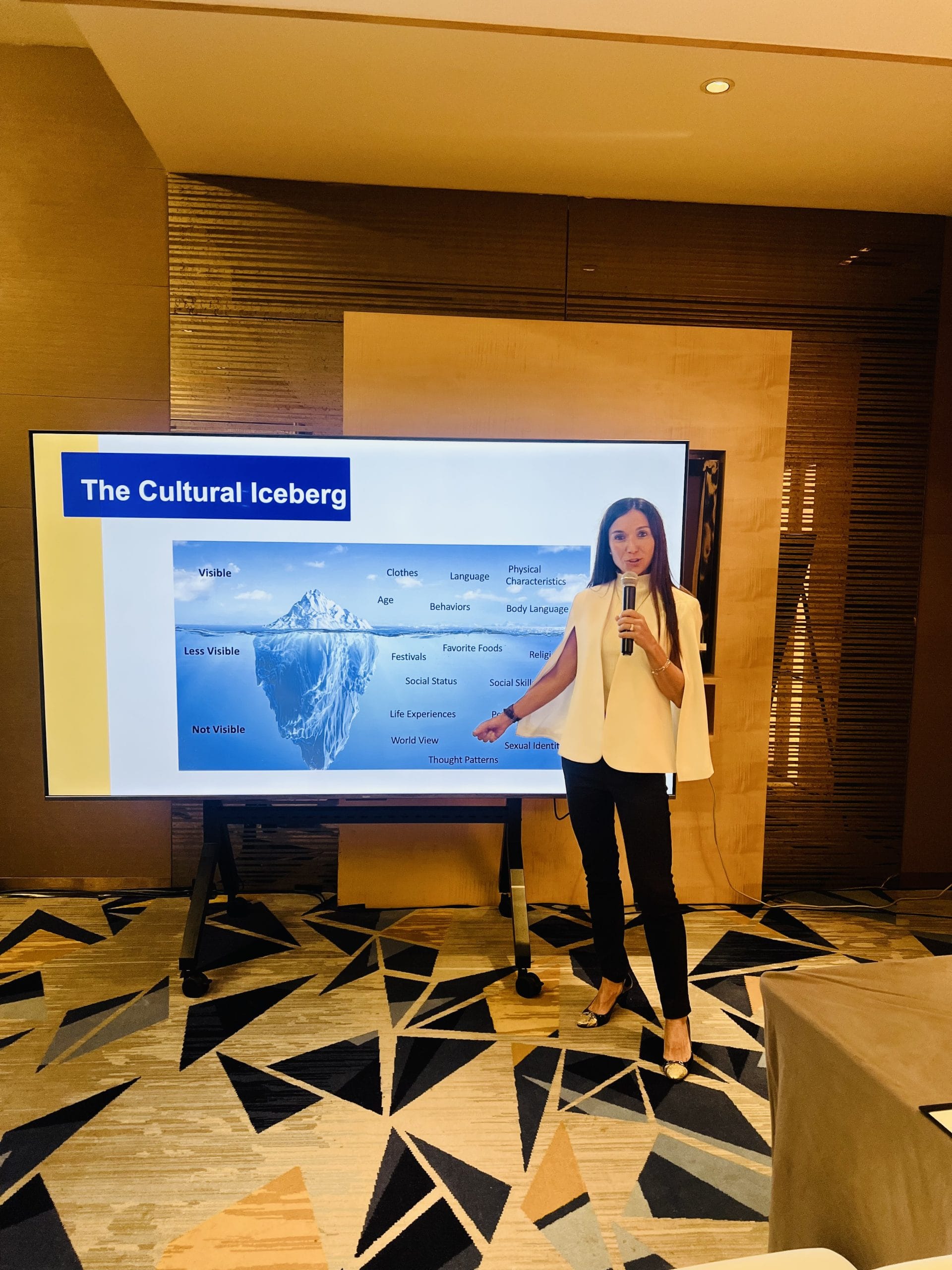 Woman with a mic delivering a presentation on cultural agility. She's wearing a white jacket and black pants.