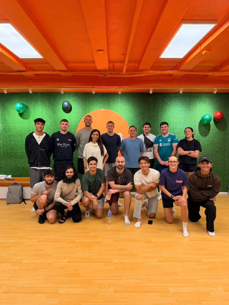 Picture of a group of people in a workshop. Mostly guys and 3 women with a green background